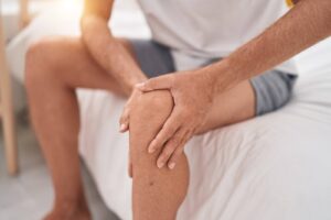 Man sitting on a bed holding his knee, appearing to experience knee pain or discomfort.